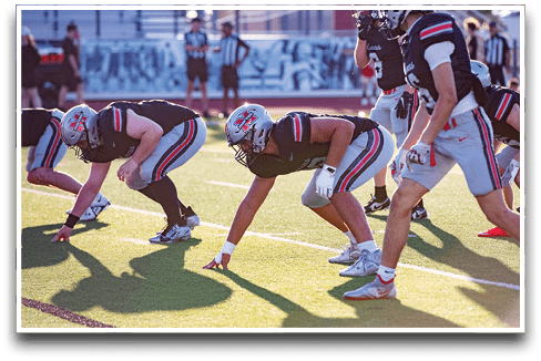 People in football uniforms are crouched in a ready position on a sunlit field preparing for a play. Description generated by AI
