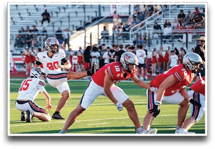 A group of people in football uniforms are on a field with one person kicking a football held by another person while others prepare for the play. Description generated by AI