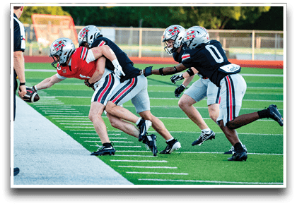 Three people in football uniforms are actively engaged in a play on a green field near the sideline, with one person reaching out to catch or hold a football while being tackled by the others. Description generated by AI