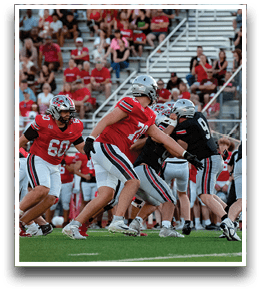 Adults wearing football helmets and uniforms are actively engaged in a game on a grassy field with spectators in the background. Description generated by AI