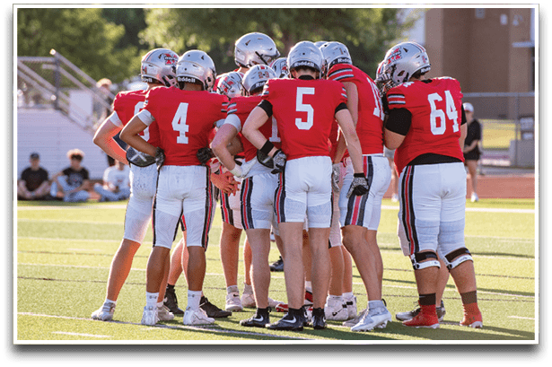 A group of people wearing red and white football uniforms and helmets are huddled together on a sunlit field. Description generated by AI