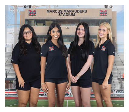 Four adults wearing matching black polo shirts and shorts stand smiling on a sports field in front of stadium bleachers and a building labeled Marcus Marauders Stadium. Description generated by AI