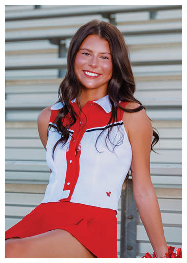 A person with long wavy hair is sitting on bleachers wearing a red and white sleeveless top with fringe and a red skirt, smiling at the camera. Description generated by AI