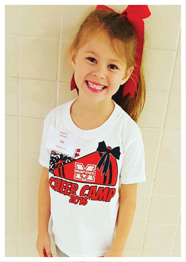 A smiling child with a ponytail tied with a large red bow is wearing a white t-shirt that says “Cheer Camp 2016" and standing against a tiled wall. Description generated by AI
