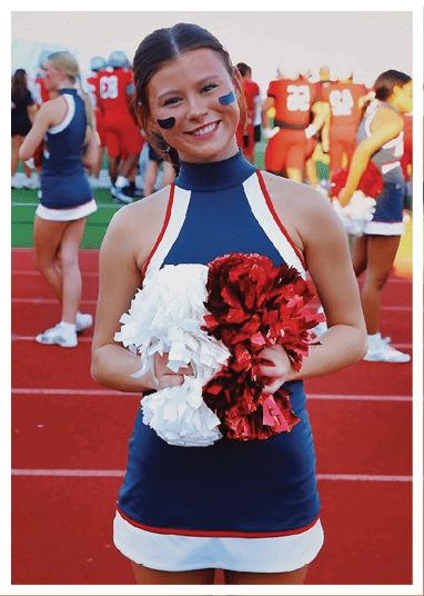 A person in a cheerleading uniform with face paint is smiling and holding red and white pom-poms on a sports field with other people and players in the background. Description generated by AI