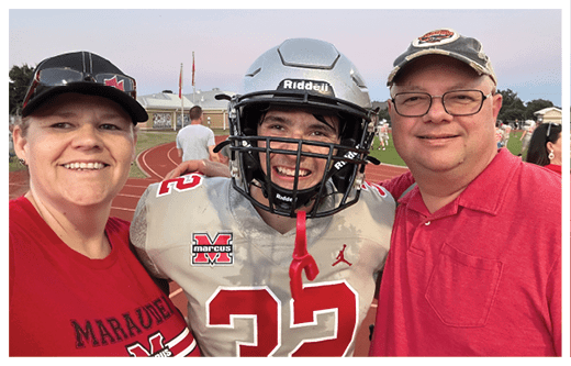Three people stand closely together on a sports field, with one person wearing a football uniform and helmet and the others wearing red shirts. Description generated by AI