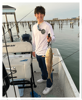A person with tousled hair wearing a white long-sleeve shirt and sunglasses hanging from the collar is holding a fishing tool with a fish attached while standing on a boat near wooden piers over calm water. Description generated by AI
