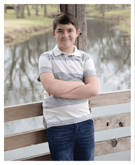 A person with short dark hair wearing a striped polo shirt is standing outdoors with arms crossed and smiling slightly in front of a wooden post near a body of water. Description generated by AI