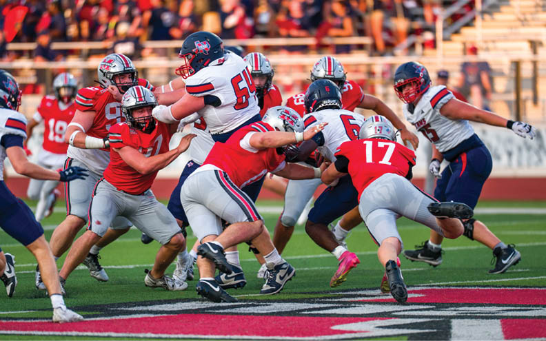 People in football uniforms are engaged in a close, intense play on a green field with a crowd in the background. Description generated by AI