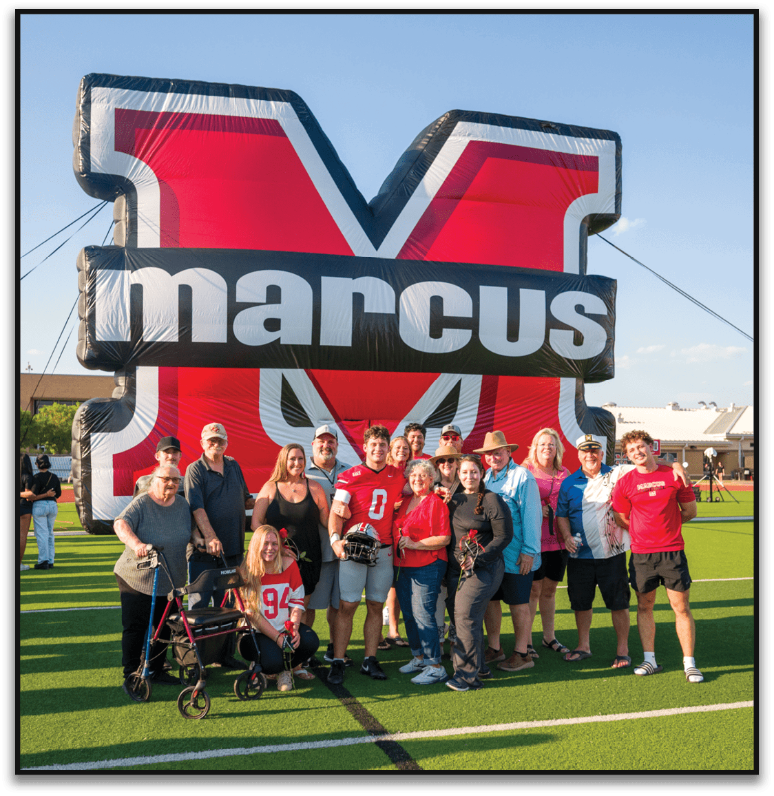 A group of people posing on a sports field in front of a large inflatable red and black letter M with the word marcus across it, with some wearing sports jerseys and one person using a walker. Description generated by AI