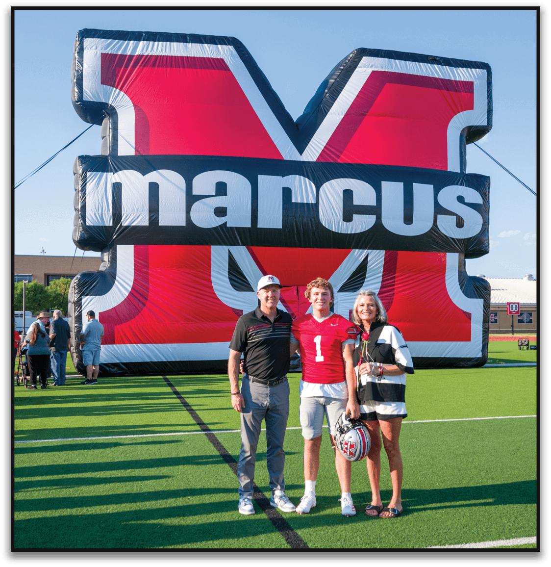 Three people stand on a football field in front of a large inflatable red and black letter M with the word marcus across it, with one person wearing a red football jersey and holding a helmet. Description generated by AI