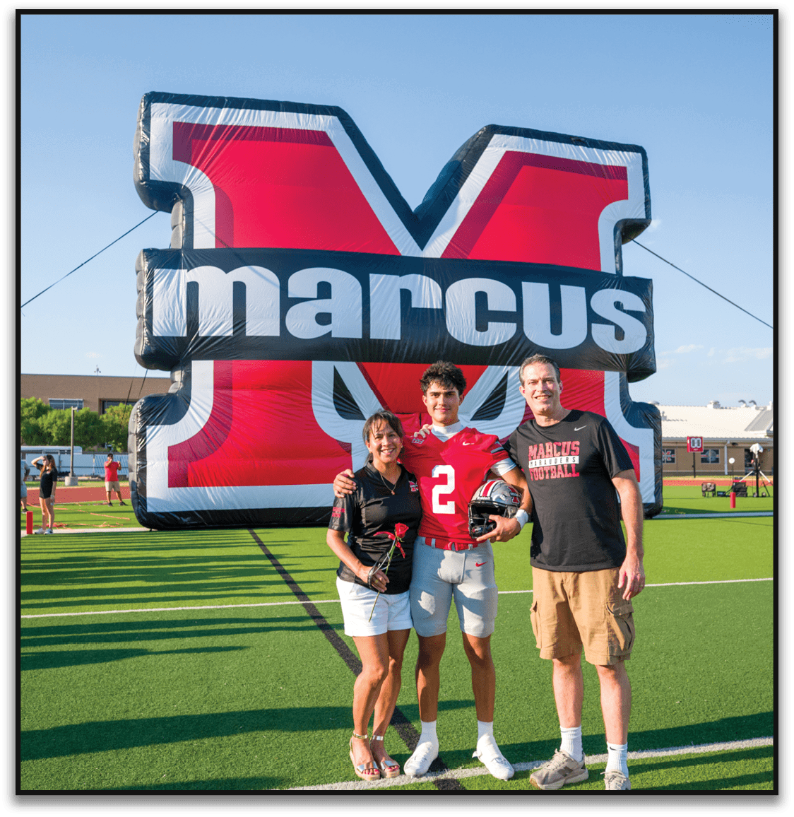 Three people stand on a football field smiling with one person in a red football uniform holding a helmet, in front of a large inflatable red and black letter M with the word marcus on it. Description generated by AI