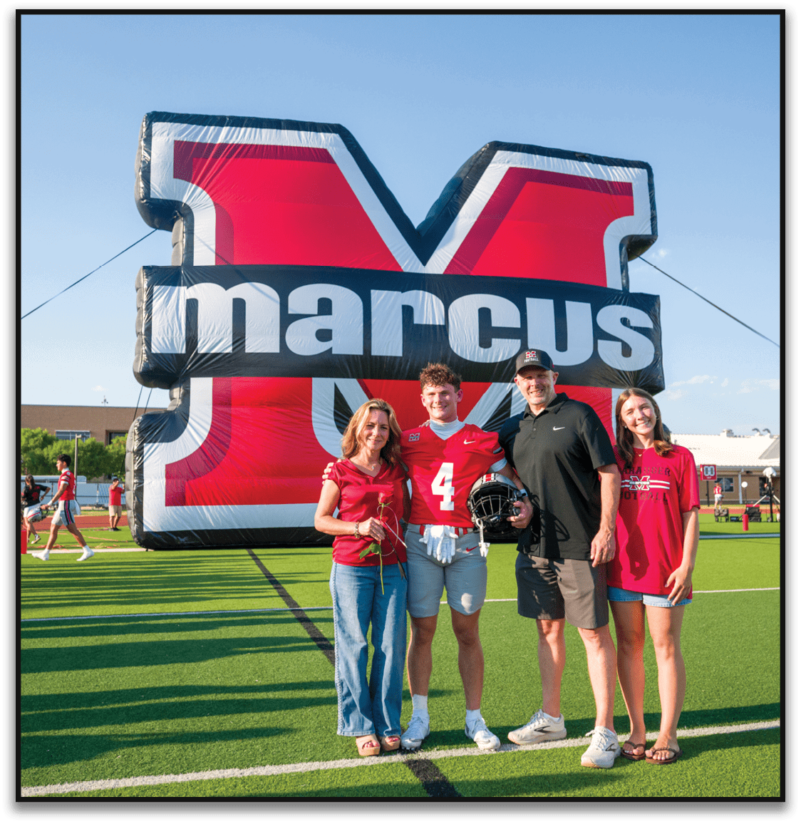 Four people stand on a football field in front of a large inflatable red and black letter M with the word marcus across it, with one person wearing a football uniform holding a helmet. Description generated by AI
