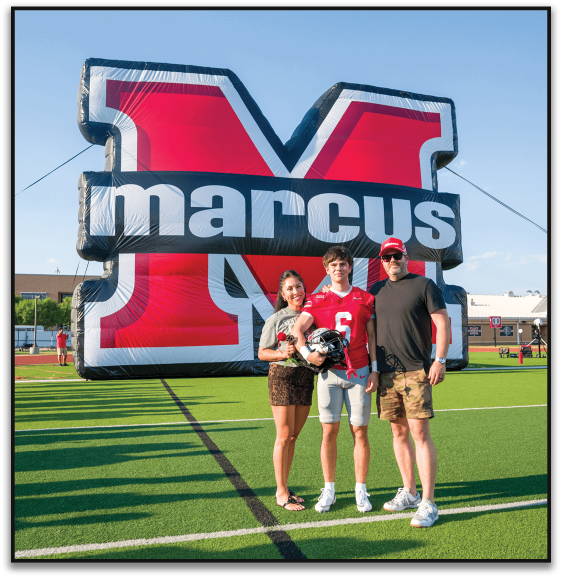 Three people stand on a football field in front of a large inflatable red and white letter M with the word marcus across it, with one person wearing a football uniform and holding a helmet. Description generated by AI