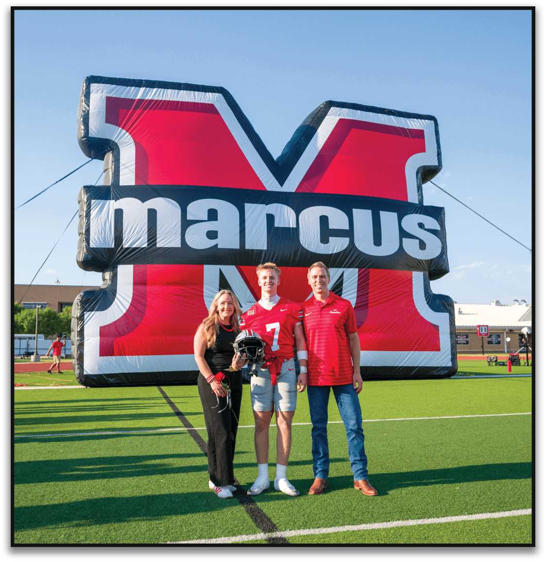 Three people stand on a football field in front of a large inflatable red and black letter M with the word marcus on it, with one person holding a football helmet. Description generated by AI