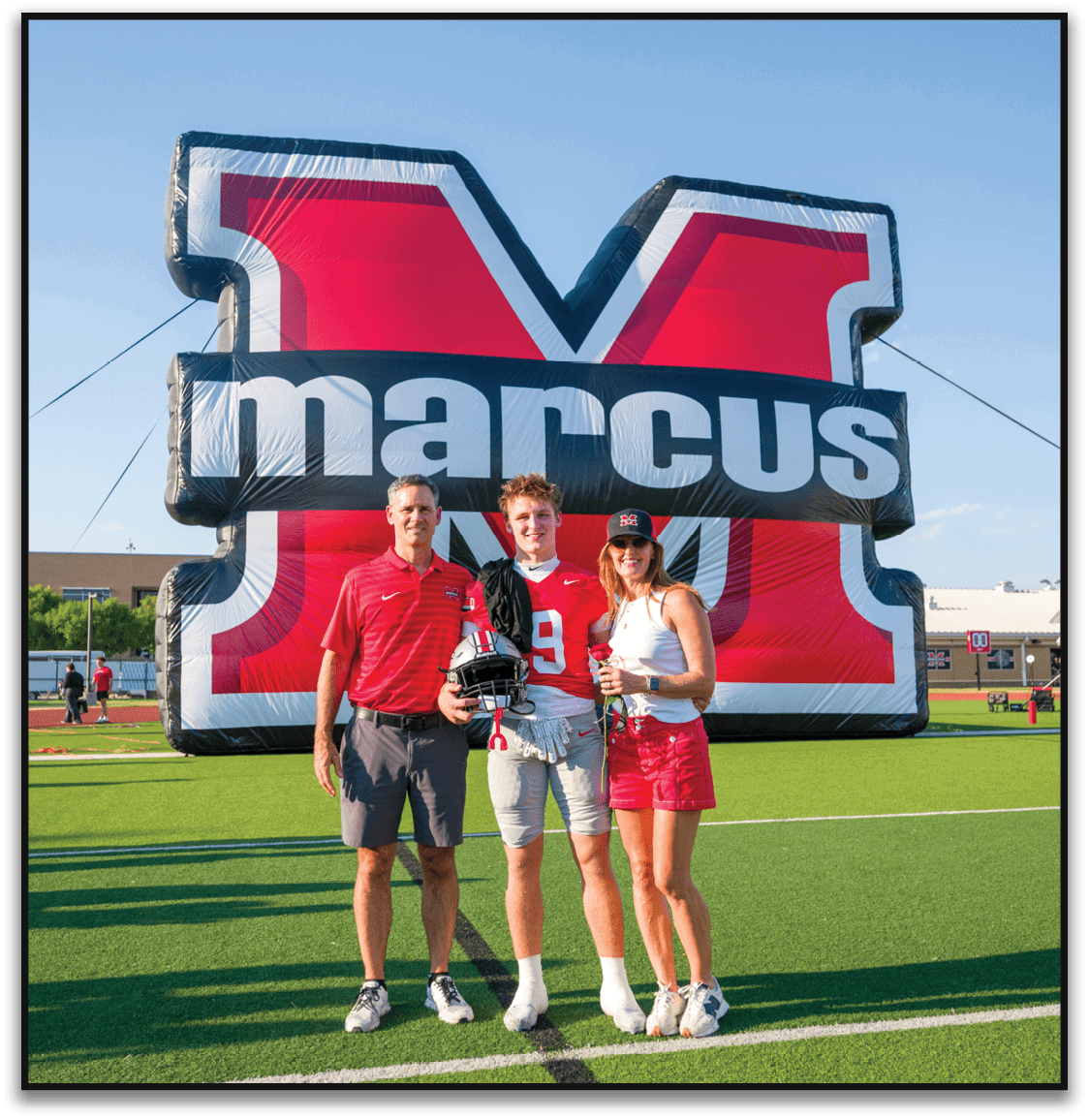 Three people stand on a football field in front of a large inflatable red and black letter M with the word marcus on it, with one person wearing a football uniform and holding a helmet. Description generated by AI