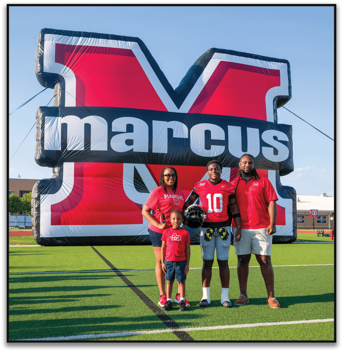 Four people standing on a football field in front of a large inflatable red and black letter M with the word marcus on it, with one person wearing a football uniform and holding a helmet. Description generated by AI