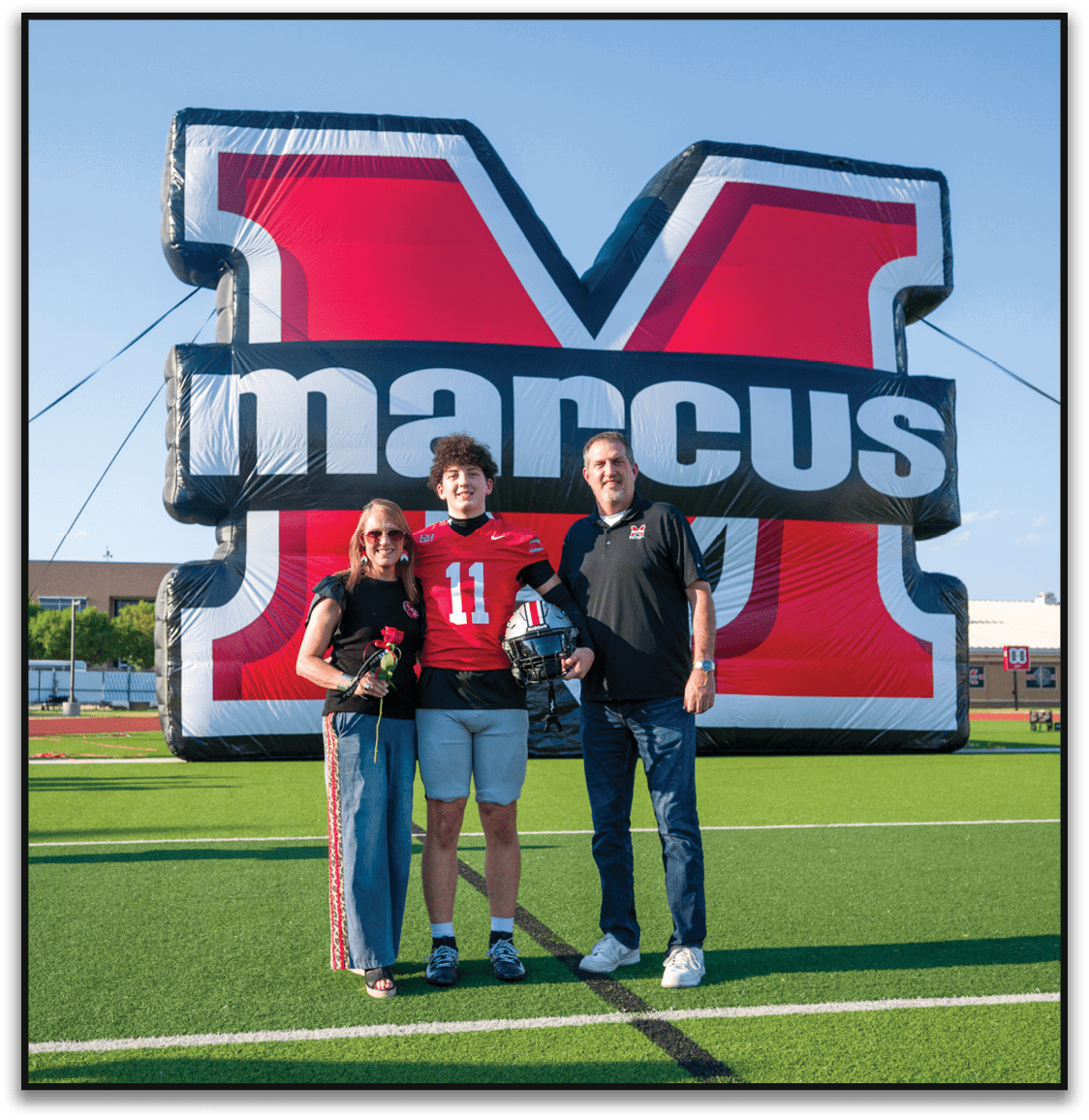 A person wearing a red sports jersey with the number 11 stands between two adults in front of a large red and black inflatable backdrop with partial text visible. Description generated by AI