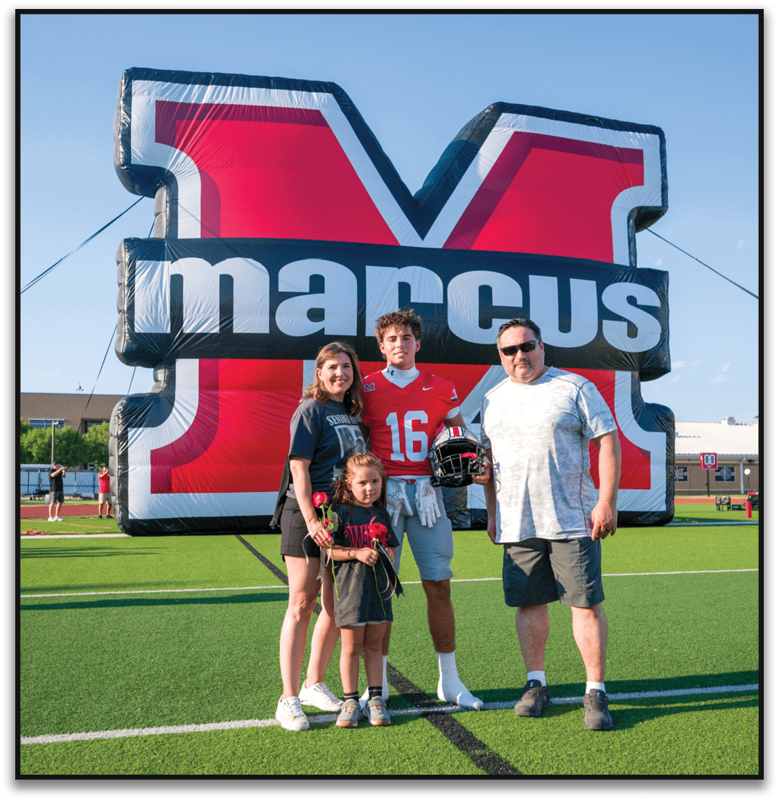 Three people stand smiling in front of a large red and black inflatable structure with white text, with one person wearing a red sports jersey and the others in casual clothing. Description generated by AI