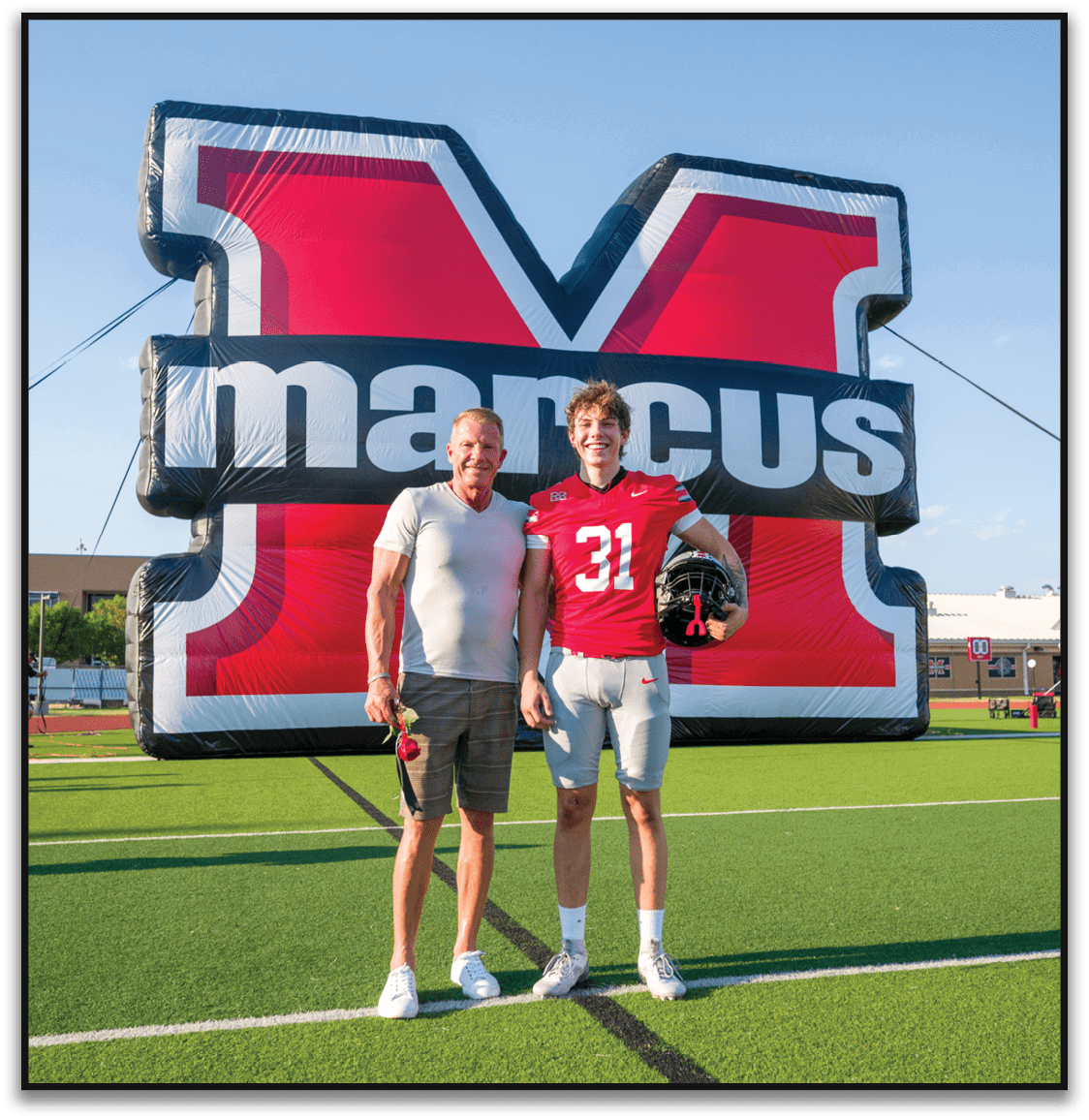 An adult and a person wearing a red football jersey with the number 31 are standing side by side in front of a large red and black inflatable backdrop, with the person in the jersey holding a football helmet and smiling. Description generated by AI