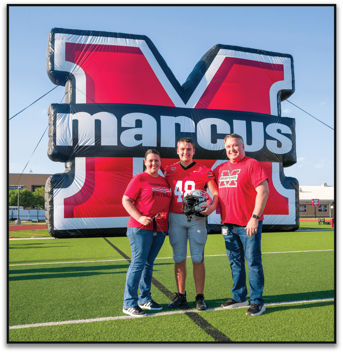 Three people wearing red Marcus football shirts stand smiling in front of a large red and black inflatable backdrop with the word Marcus partially visible. Description generated by AI