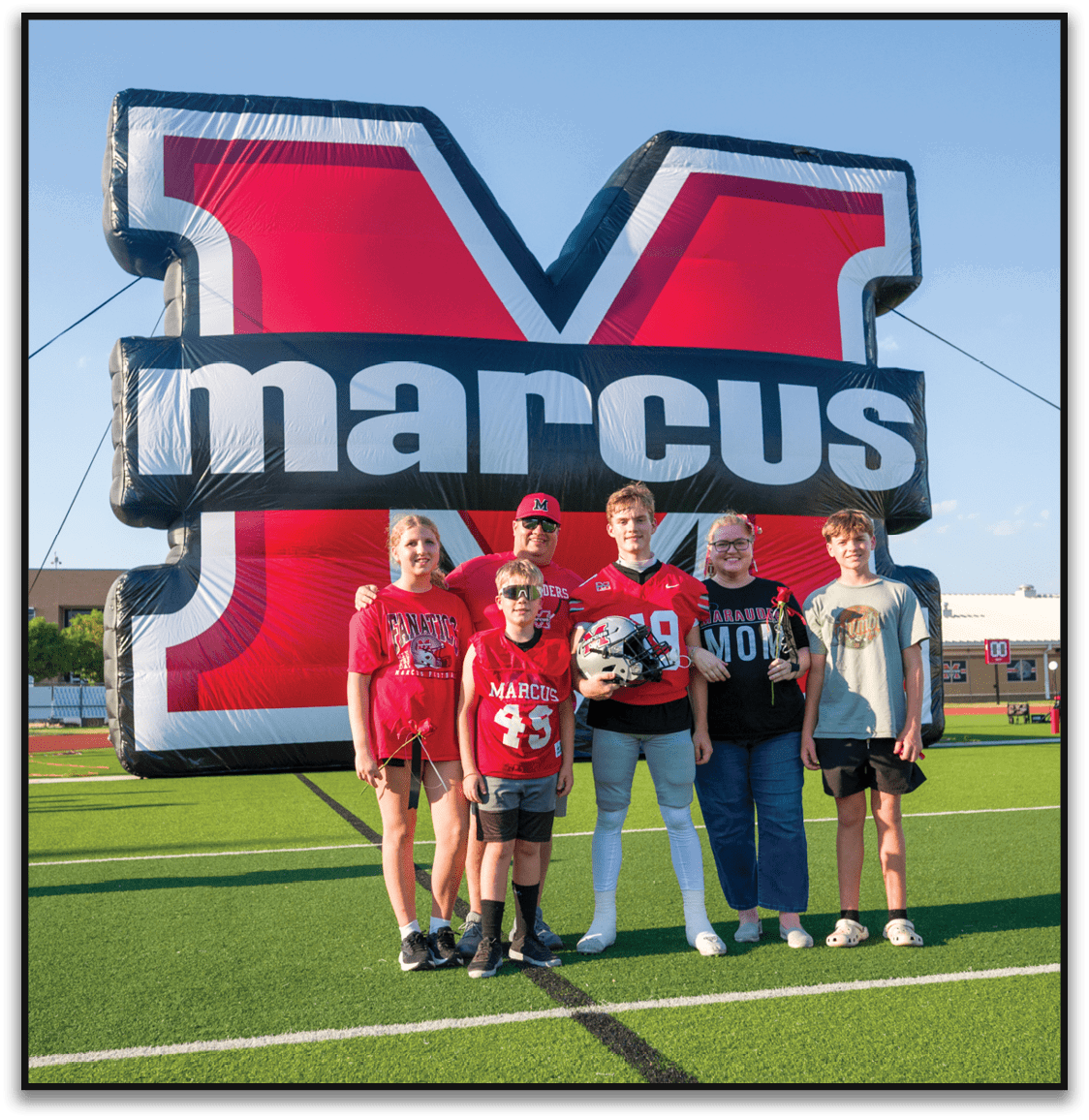 A group of five people wearing sports jerseys and casual clothing pose smiling in front of a large inflatable structure with bold text. Description generated by AI