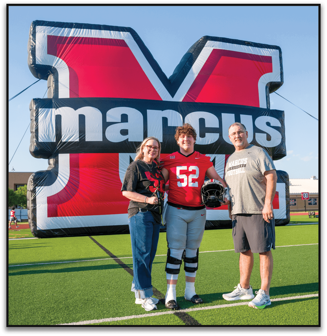 Three people smiling and posing in front of a large inflatable structure with bold red, black, and white colors and text, with one person wearing a red sports jersey. Description generated by AI