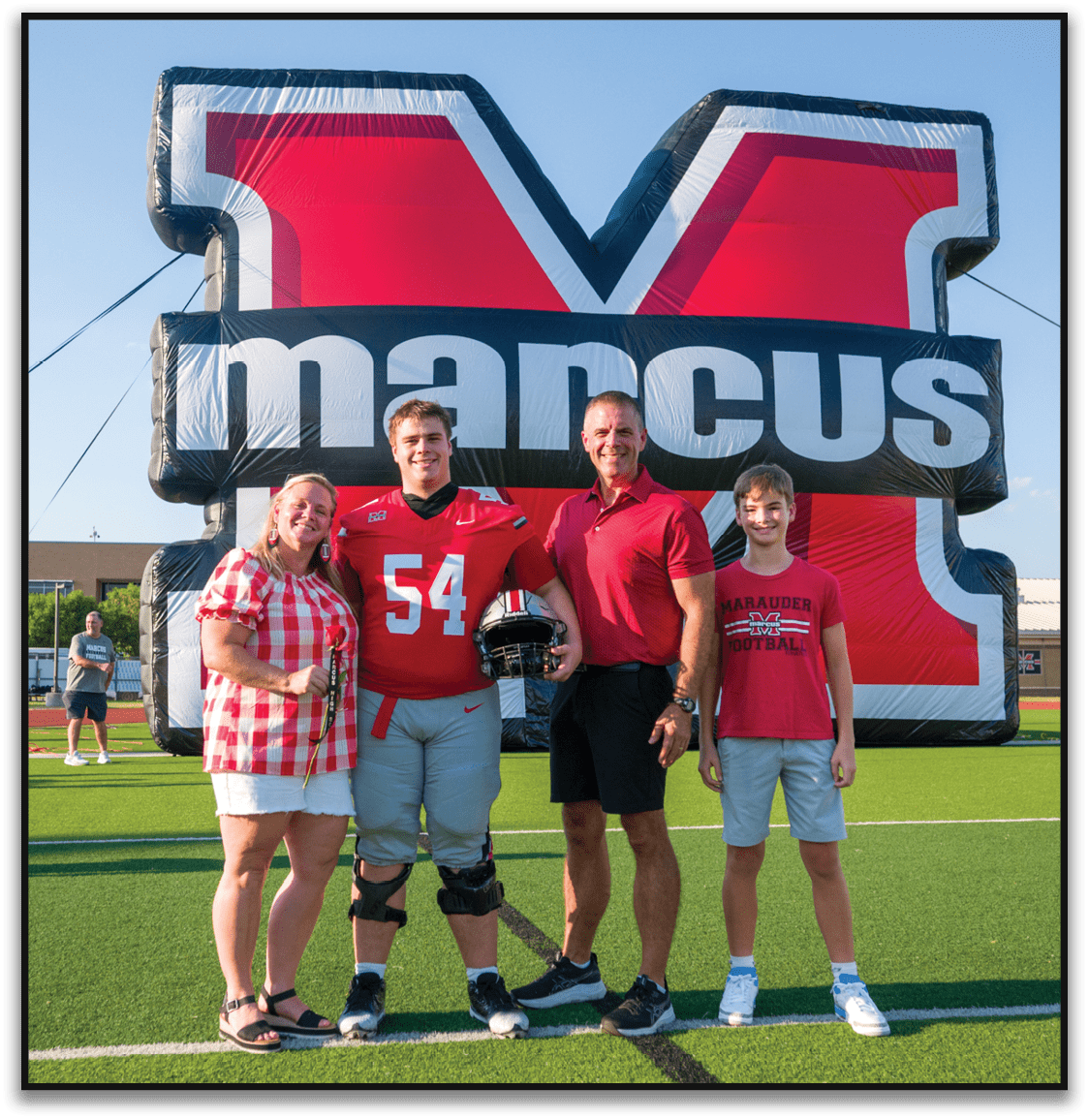 Three people stand smiling in front of a large red and black banner, with one person wearing a red sports jersey holding a football helmet and another wearing a red shirt with “Marauder Football" printed on it. Description generated by AI