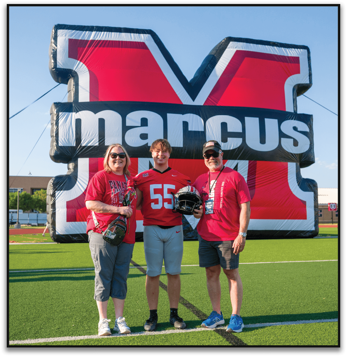 Three people wearing red shirts and sunglasses stand smiling in front of a large inflatable backdrop with bold red, white, and black colors and partially visible text. Description generated by AI