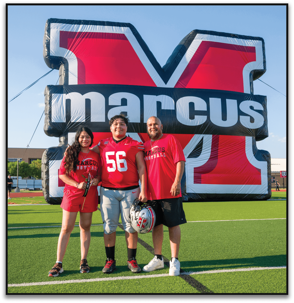 Two people wearing red Marcus football shirts stand smiling in front of a large inflatable Marcus logo. Description generated by AI