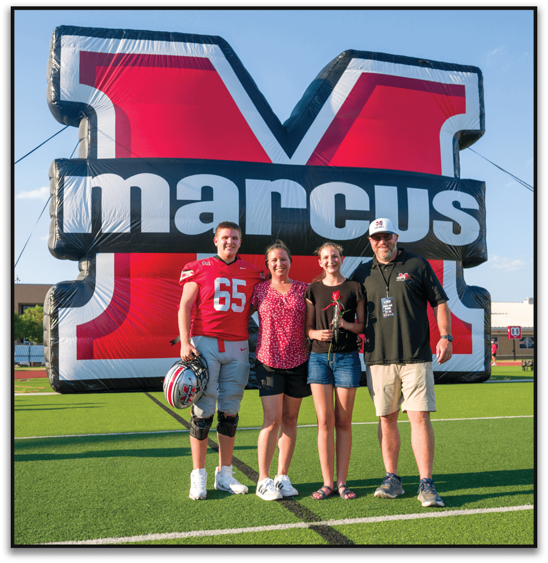 Four people smiling and standing close together in front of a large red and black inflatable backdrop with partial white text, with one person wearing a red sports jersey and another wearing a white cap. Description generated by AI