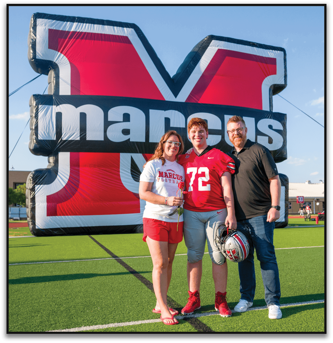 Three smiling adults stand closely together in front of a large inflatable structure with bold red, white, and black colors, with one person wearing a red sports jersey numbered 72. Description generated by AI