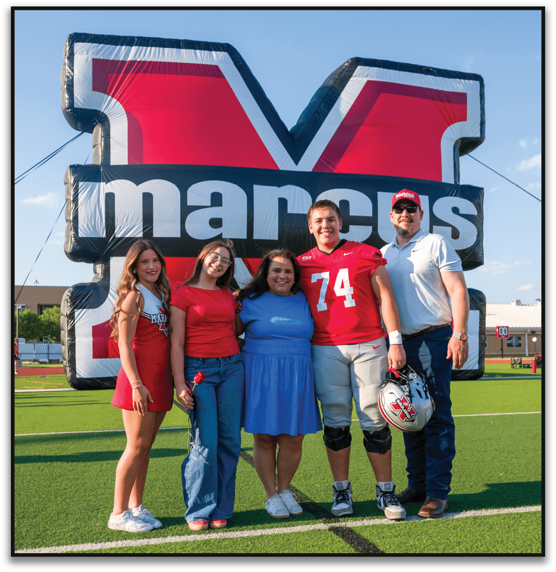 Three people smiling and posing in front of a large inflatable sign with one person wearing a red sports jersey and another wearing a red cap and sunglasses. Description generated by AI
