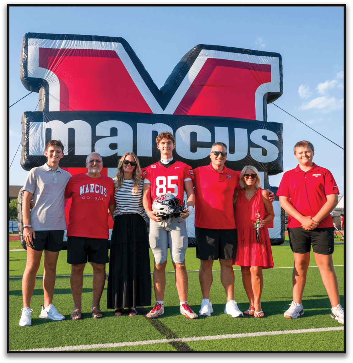 Three people wearing red shirts stand smiling in front of a large inflatable sign with bold letters and a blue sky background. Description generated by AI