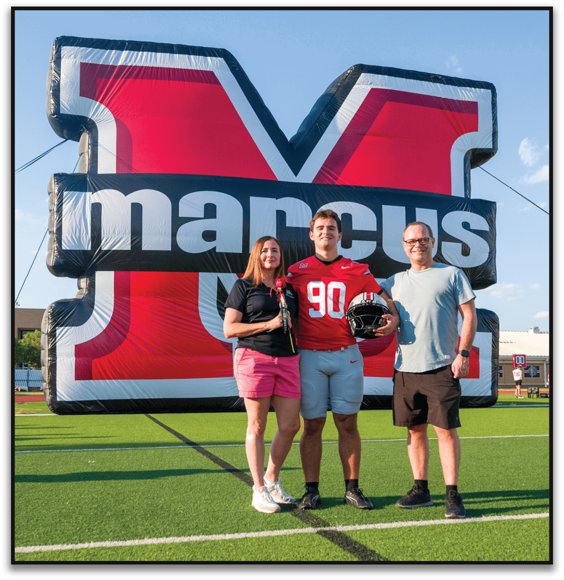 Three people stand smiling in front of a large inflatable structure with bold red, black, and white colors, with one person wearing a red sports jersey holding a football helmet. Description generated by AI