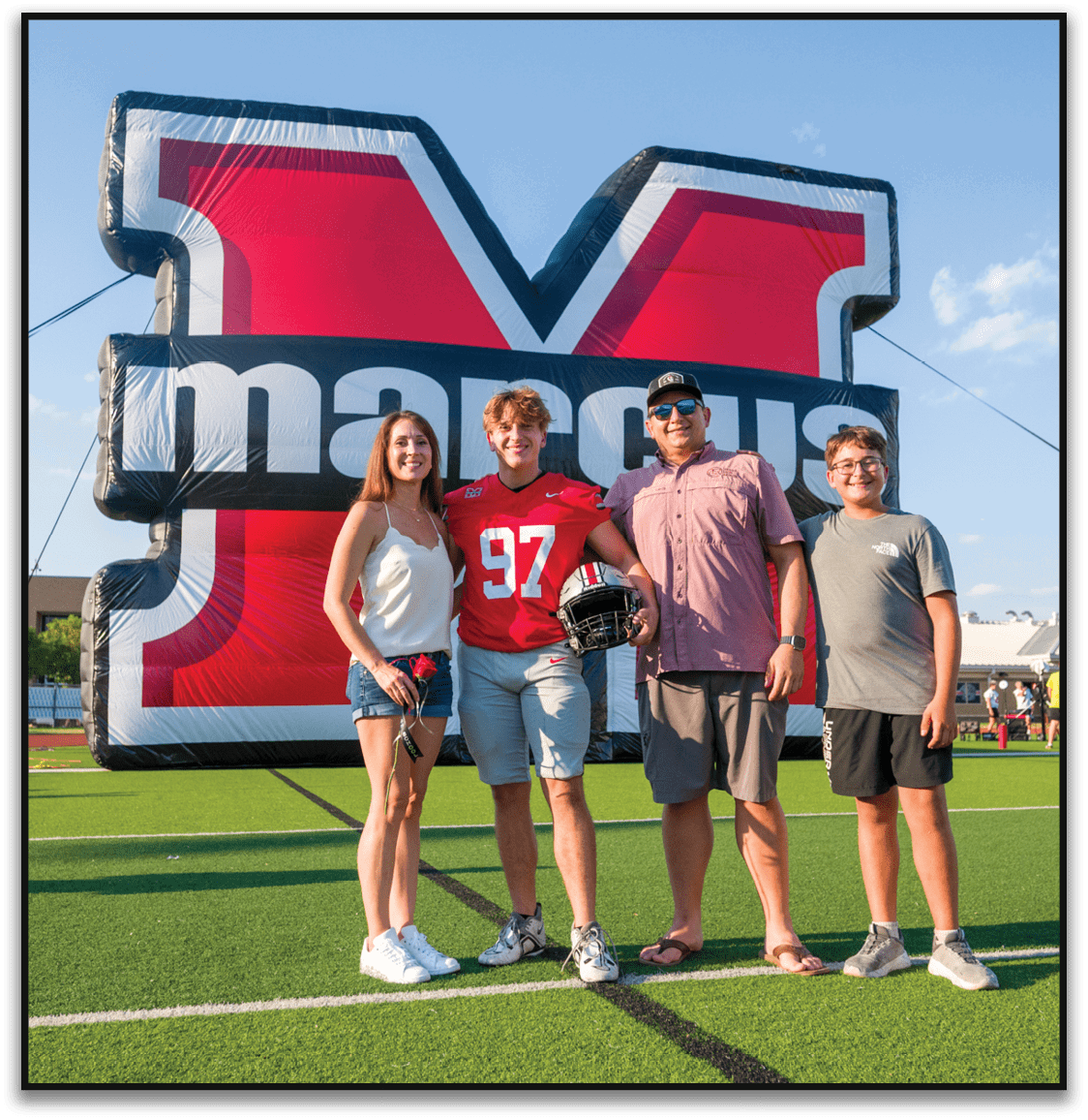 Three people stand smiling in front of a large inflatable structure with bold red, white, and black colors, one wearing a red sports jersey holding a football helmet. Description generated by AI
