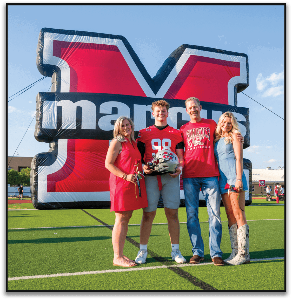 Three people stand smiling in front of a large inflatable sports logo, with one person wearing a red football jersey and holding a helmet, another wearing a red shirt with a football graphic, and the third wearing a light blue dress. Description generated by AI