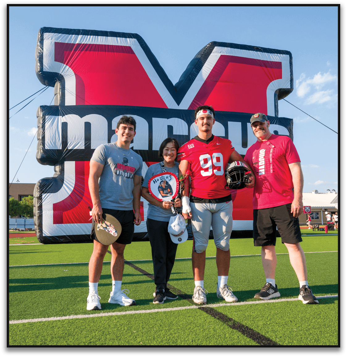 Three people stand smiling in front of a large inflatable sports-themed backdrop, with one person wearing a red football jersey holding a helmet and another holding a circular sign with a photo and the word “HOSKINS." Description generated by AI