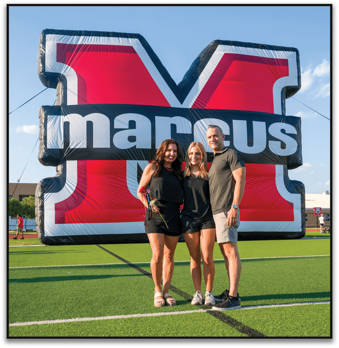 Three adults standing closely together smiling in front of a large inflatable sign with bold red, black, and white colors. Description generated by AI