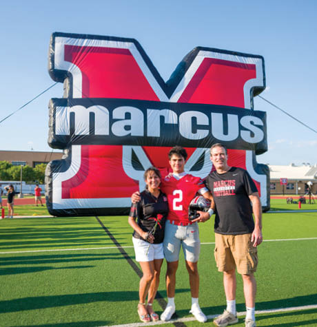 Three people stand on a football field in front of a large inflatable red and black letter M with the word marcus on it, with one person wearing a red football uniform holding a helmet. Description generated by AI