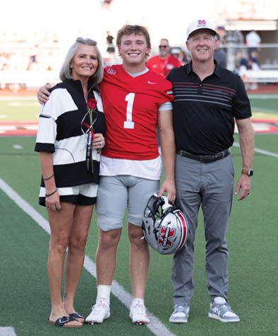 A person in a red football uniform stands on a field holding a helmet and a rose, flanked by two adults smiling and posing for a photo. Description generated by AI