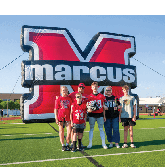 Six people standing on a football field in front of an inflatable arch with the word “marcus," with one person wearing a football uniform holding a helmet. Description generated by AI
