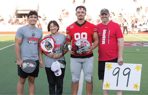 Four people stand on a football field with one person in a red football uniform holding a helmet, while others hold signs and flowers, smiling toward the camera. Description generated by AI