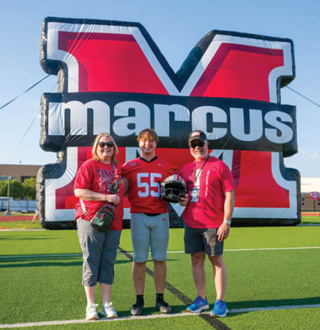Three people stand on a football field in front of a large inflatable red and black letter M with the word marcus, with one person wearing a football uniform and holding a helmet while the others wear casual clothing and sunglasses. Description generated by AI