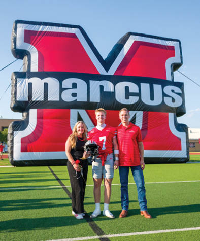 Three people stand on a football field in front of a large inflatable red and black letter M with the word marcus across it, with one person wearing a football uniform holding a helmet. Description generated by AI