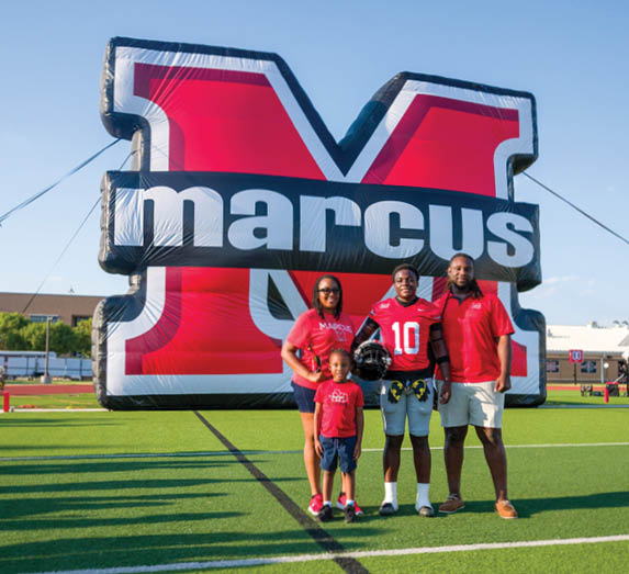 Four people including one wearing a football uniform and helmet stand on a sports field in front of a large inflatable structure with the word marcus on it. Description generated by AI