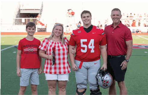Four people stand on a football field with one person wearing a red football uniform holding a helmet, while the others wear casual red and white clothing and smile at the camera. Description generated by AI