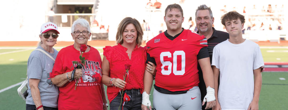 Six people stand together on a sports field, with one person wearing a red football uniform and others holding roses, all smiling and posing for the photo. Description generated by AI