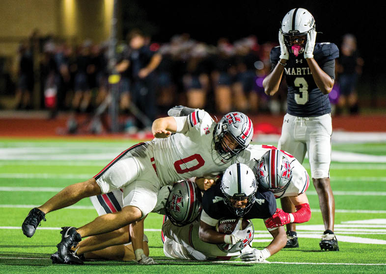 Several people in football uniforms are on a field with one person holding a football while others are tackling or standing nearby. Description generated by AI
