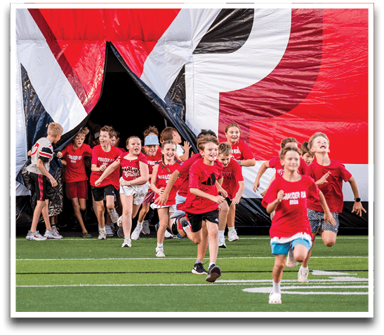 Children wearing red shirts are running excitedly out of a large inflatable tunnel onto a sports field. Description generated by AI